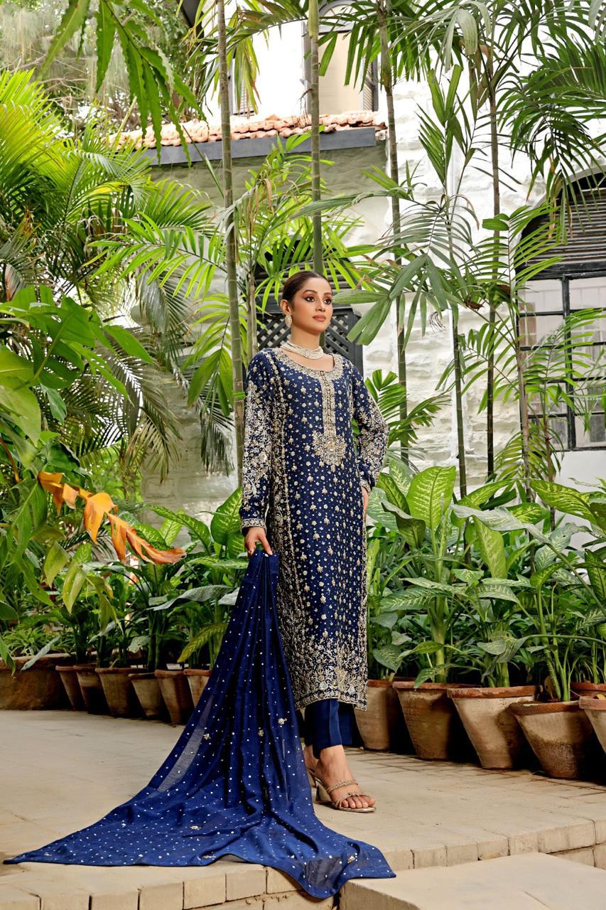 A woman standing outdoors wearing a traditional formal luxury outfit with a dupatta, primarily in navy blue color with gold details.